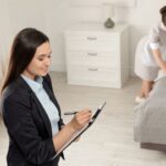 Household manager checking a task list while a housekeeper prepares a bedroom.